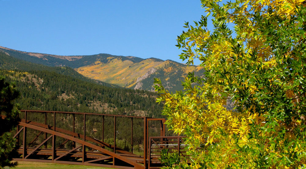 A pedestrian metal bridge sits on the left of the image, to the right yellow and green leaves are on a tree. In the distance blue sky meets mountains that are slowly becoming covered in golden hues.