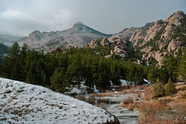 A small creek is frozen over with ice near Bailey, Colorado. A thick stand of pine trees are on the far bank with rocky peaks rising up behind them.