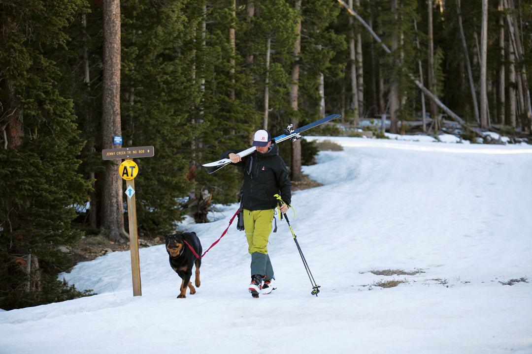 A man with carrying skis across his shoulder walks down a snowy path with a dog on a leash
