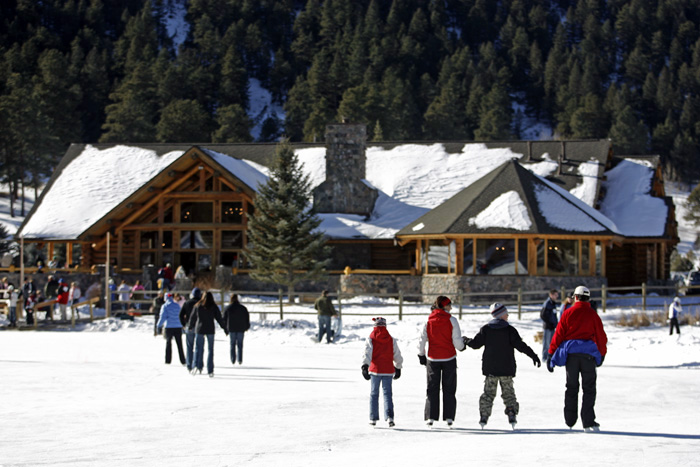 In the foreground, a family of four on ice skats holds hands as they glide toward a wooden lodge with snow on the roof. Other skaters can be seen in the background.