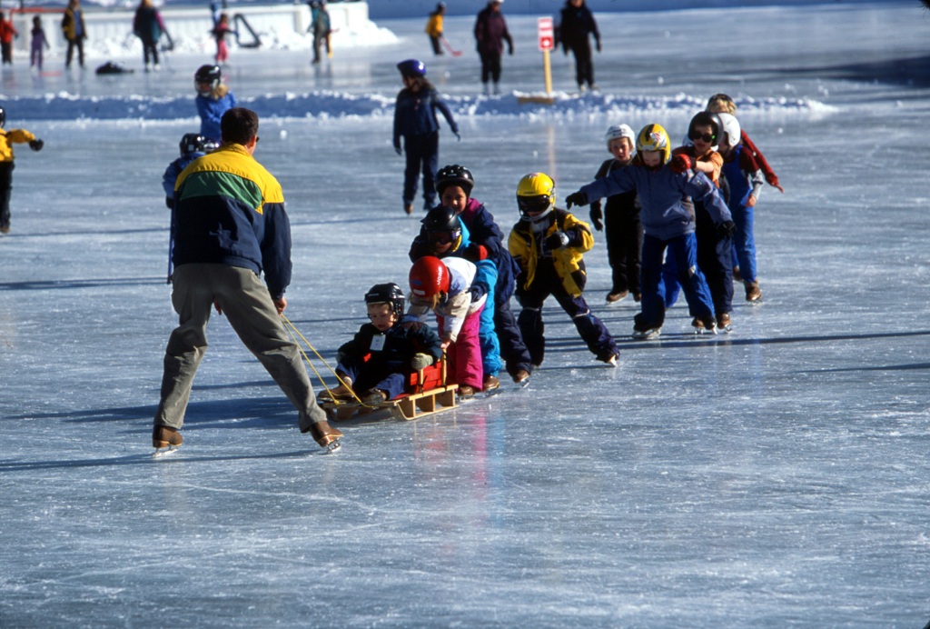 An adult pulls a kid on a sled over an ice-skating rink at the frozen Keystone Lake. Behind the sled is a line of small children learning to skate.