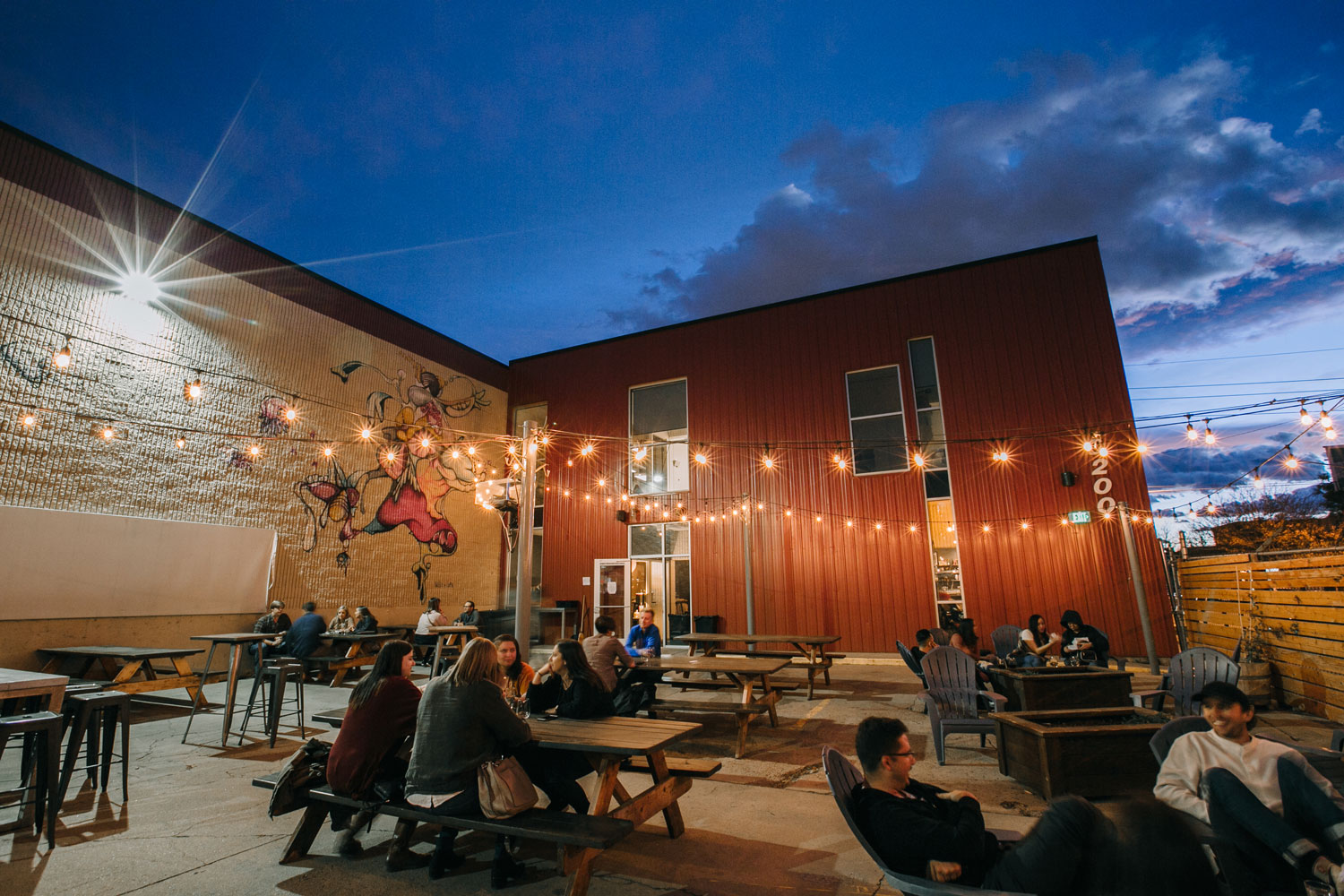 Adults sit with friends and family at wooden picnic tables and around firepits as strings of exposed-bulb lights glow overhead in Denver, Colorado.
