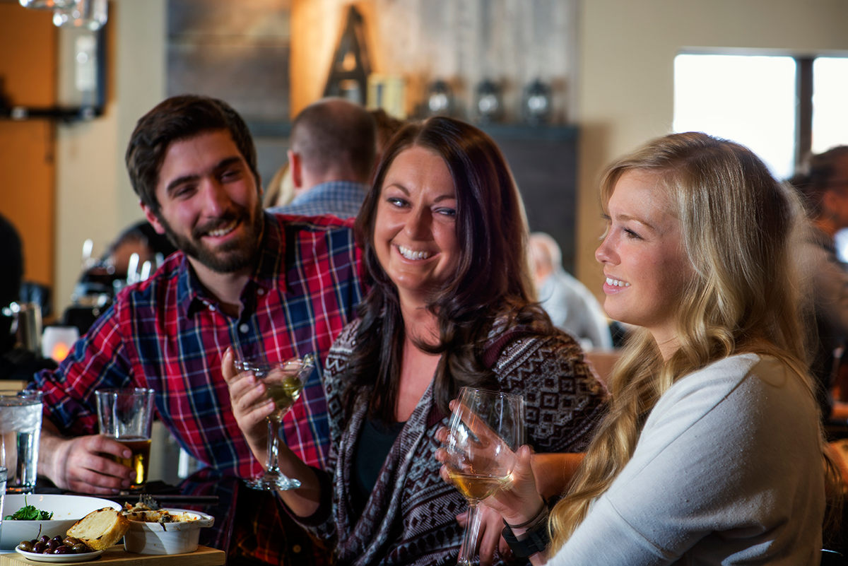 Three people laugh together holding drinks at a bar during cocktail hour at Aurum in Steamboat Springs