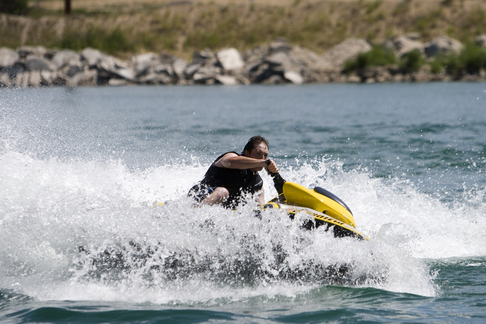 A person sits on a yellow jet ski in a large white wave on Jackson Lake. On the shore behind them there are big rocks and boulders that meet greenery.