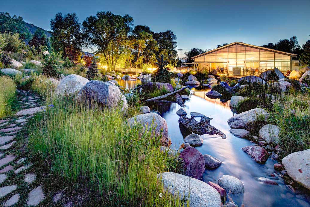 A stony walking path passes by a calm pond featuring tall grasses, boulders and fallen tree branches at the John Denver Sanctuary in Aspen, Colorado. Across the pond, a see-through building is lit up and hosts a large gathering.
