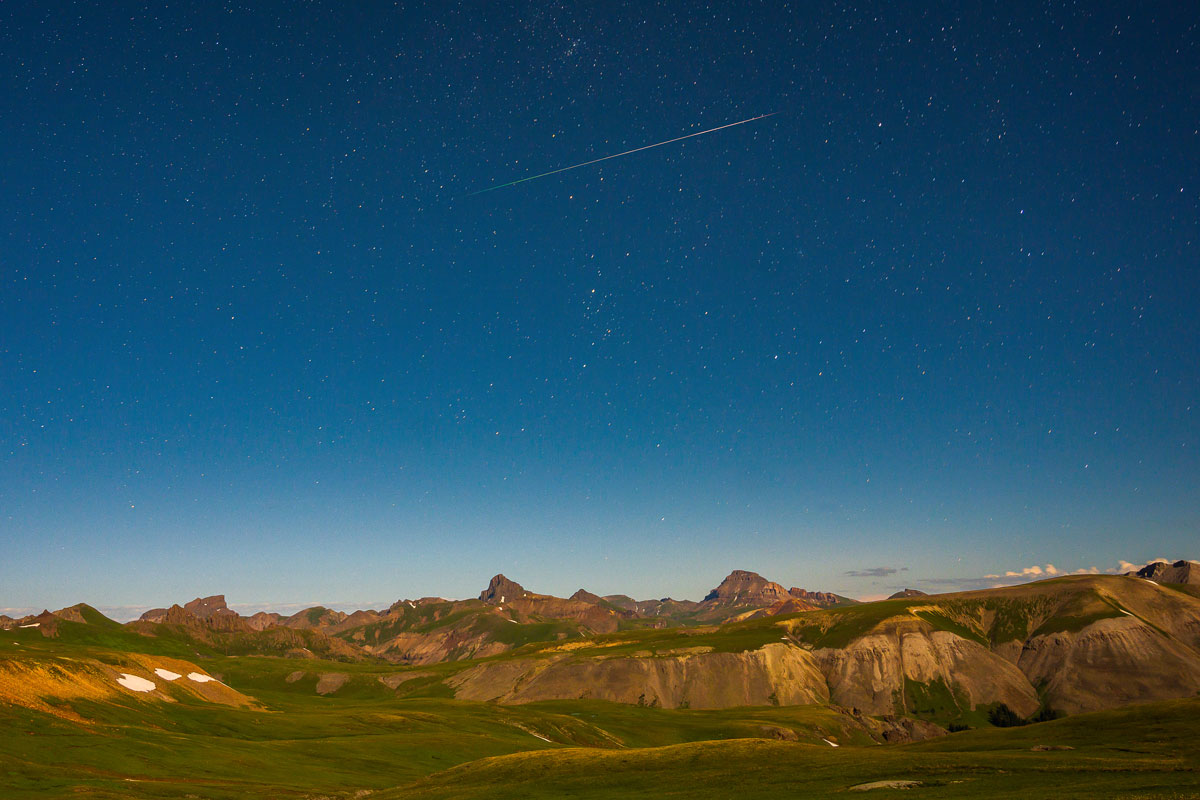 A star-filled sky at dusk sits above green fields with Rocky Mountains.