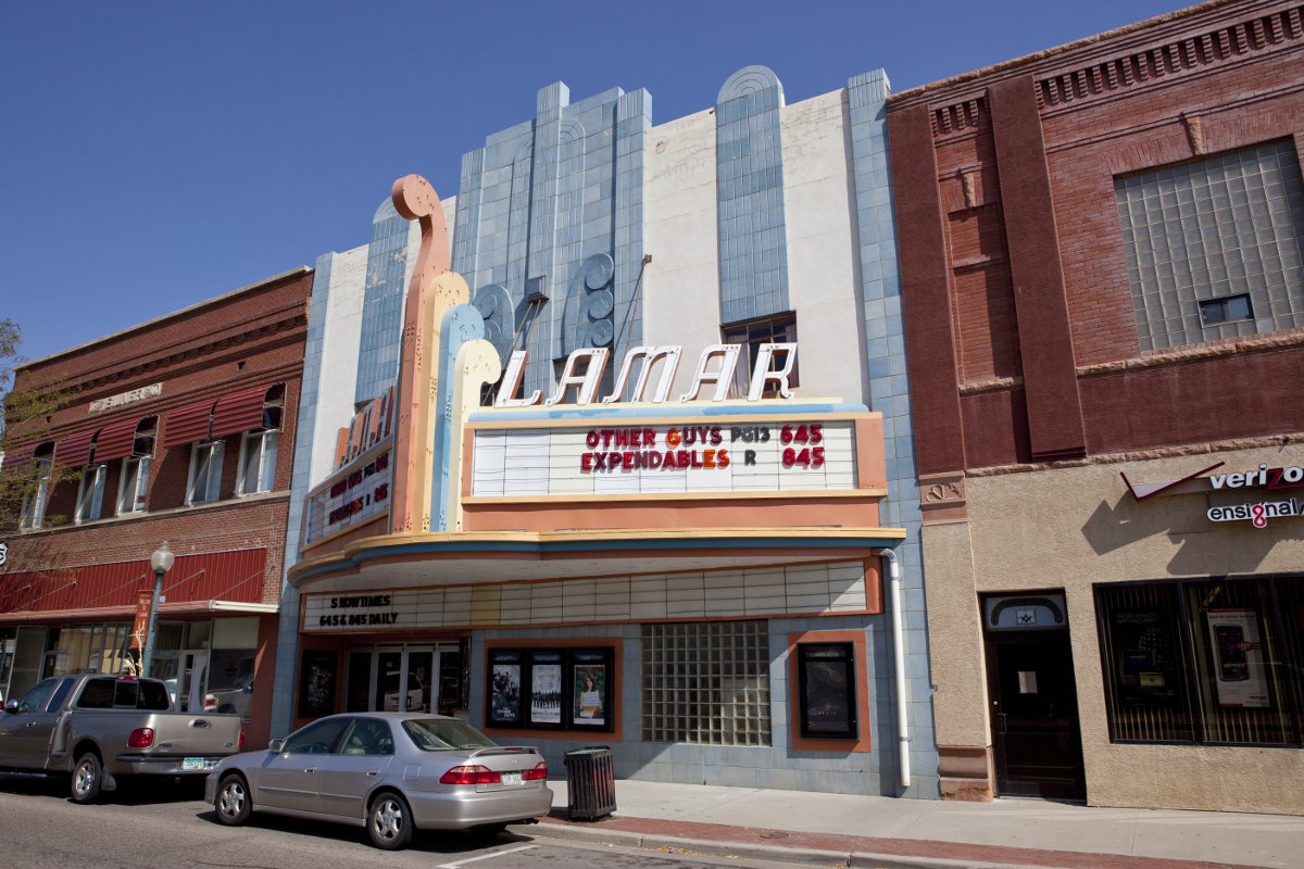 The Art Deco Lamar Theater sandwiched in between red-brick buildings. The theater is painted white, blue and orange. The marquee says "Other Guys PG13 545 Expendables R 645."