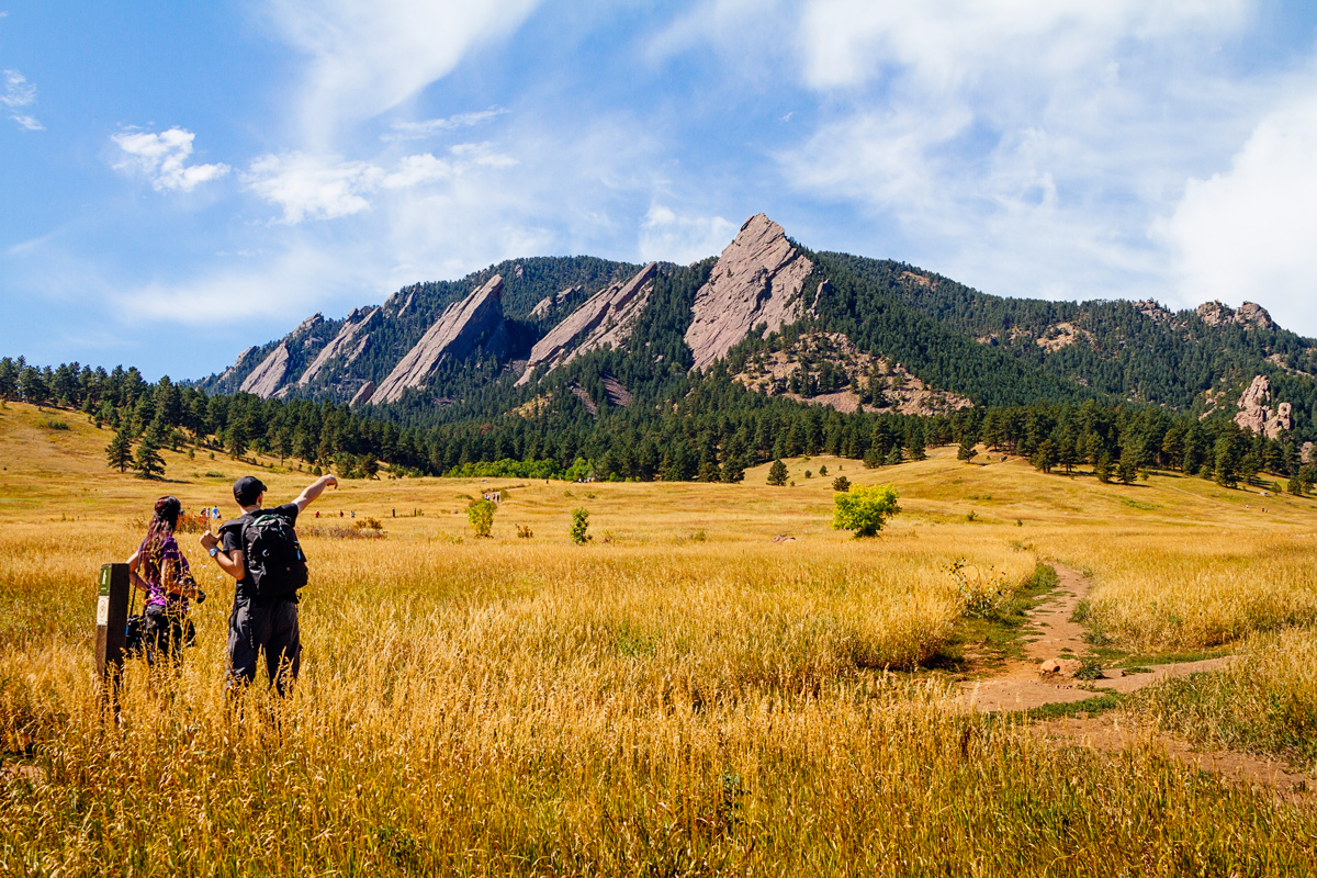Tall prairie grasses shine a deep gold beneath the late-summer sun. Several dirt paths wind through the grasses. Two hikers discuss the path leading to the tall, rocky peaks in the distance.