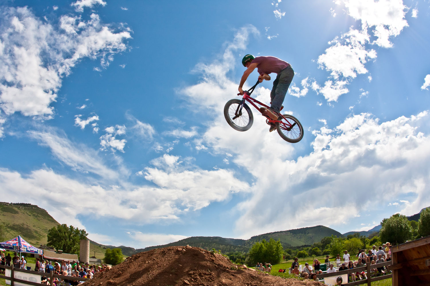 A person gets air on a bicycle against a blue sky with white clouds above a pile of red dirt. Spectators watch on in the sunshine with rolling hills to their backs.
