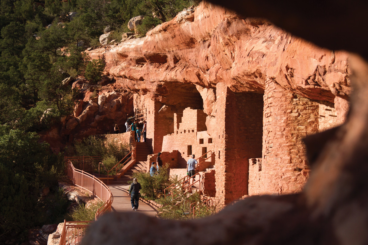 A paved walkway with a railing connects to several ancestral structures at the Manitou Cliff Dwellings. People walk the path and climb stairs to explore the dwellings' interiors.