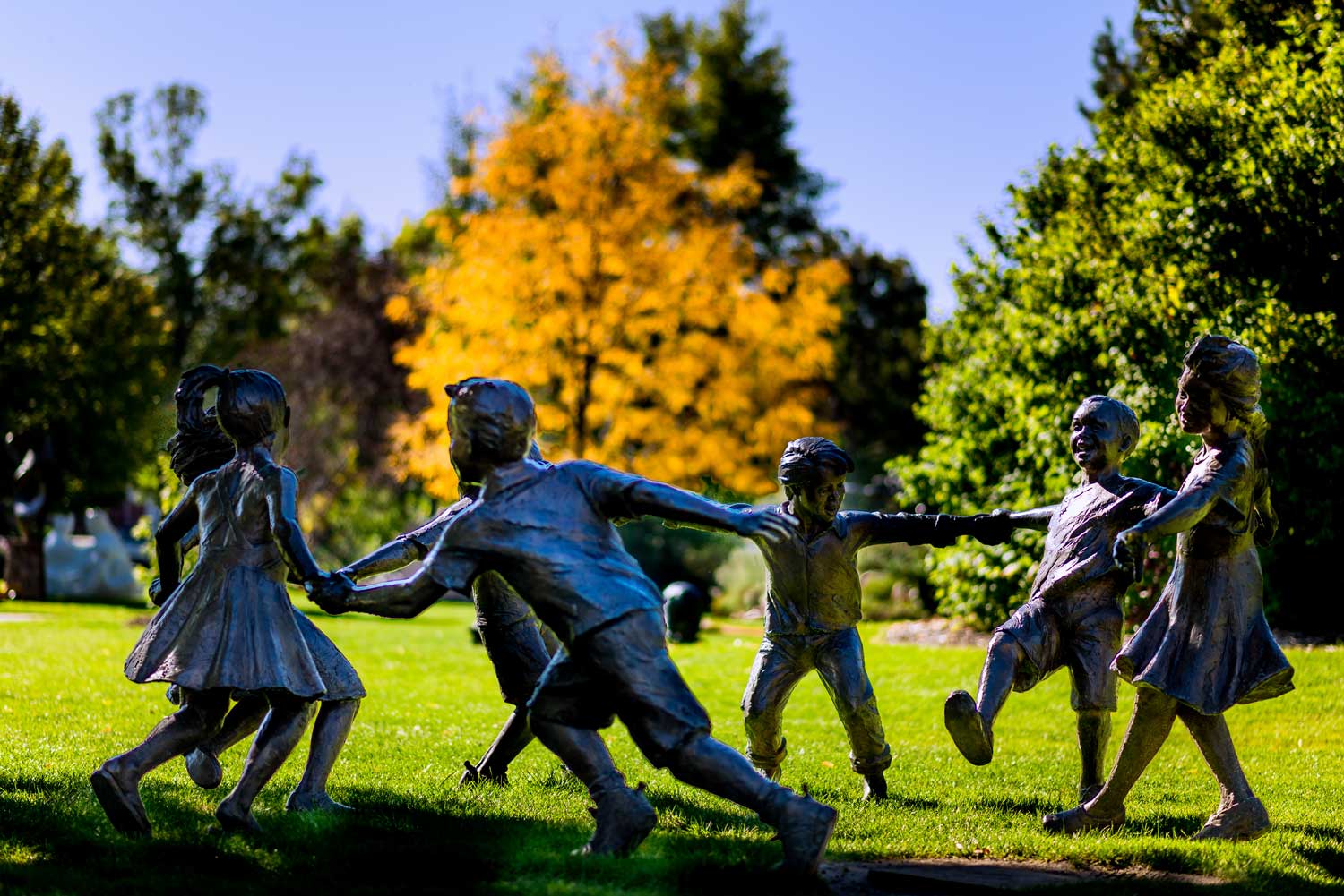 It's fall in Loveland, Colorado, and a tree in the background has bright orange-yellow foliage. In the foreground, a sculpture features several children happily playing and trying to run hand-in-hand in a circle.