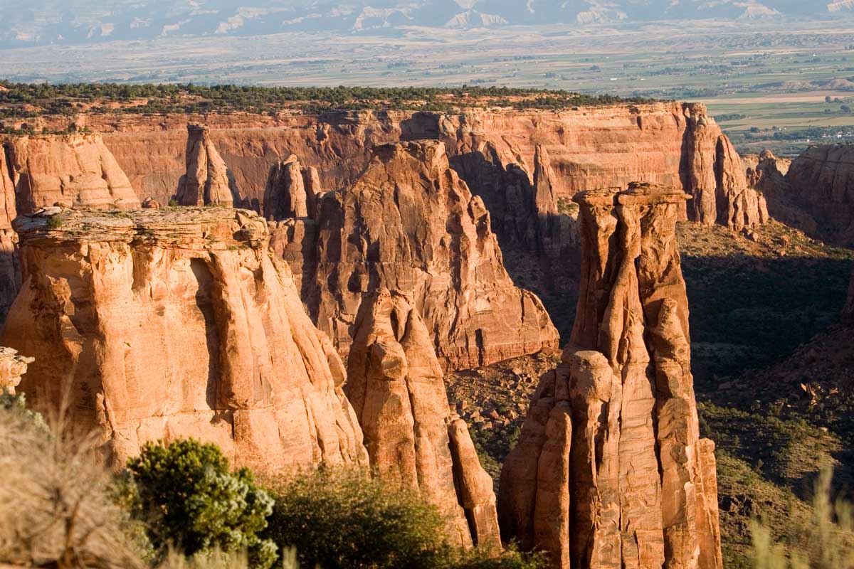 The red-rock geologic formations rise up from the green scrubby plants on land.