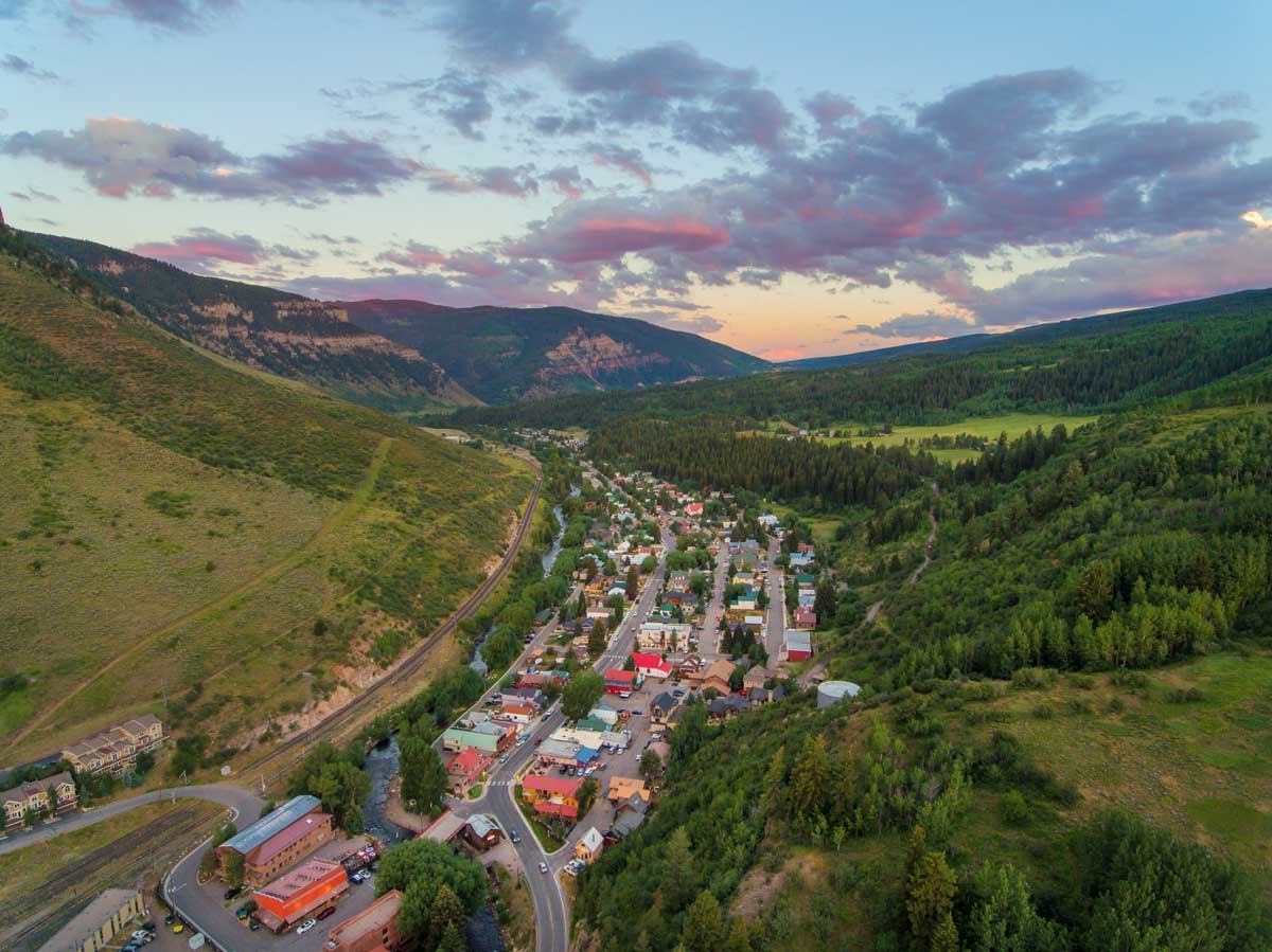 Aerial view of Minturn in the summer with rolling green hills and mountains in the distance with pink clouds above the town at dusk.