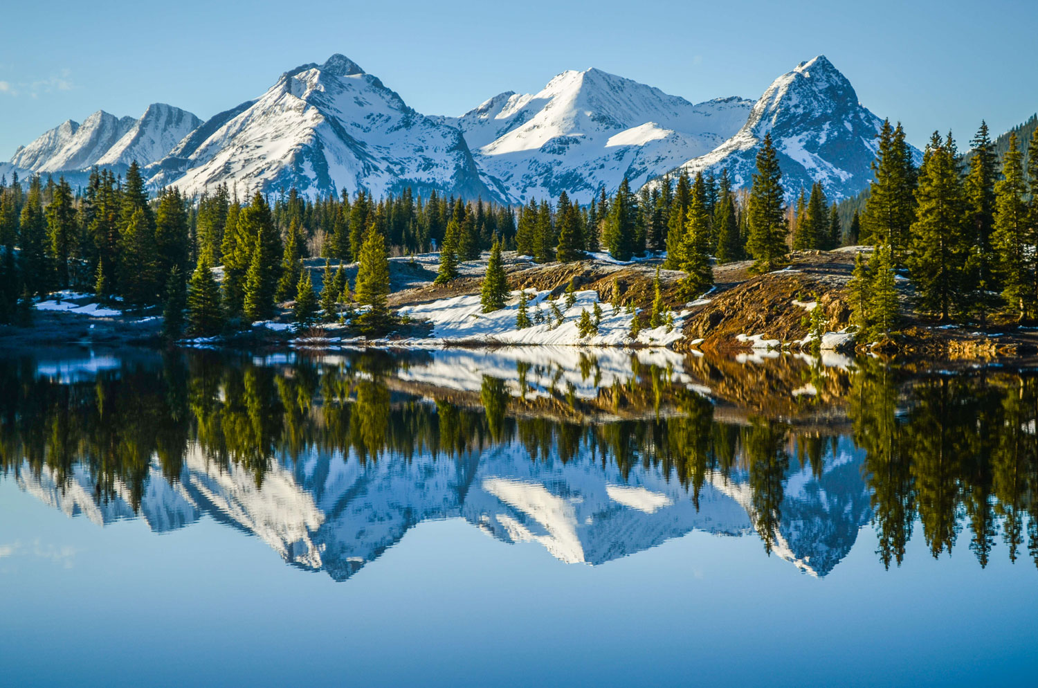 A very still lake reflects snowcapped peaks and evergreen trees on the shore