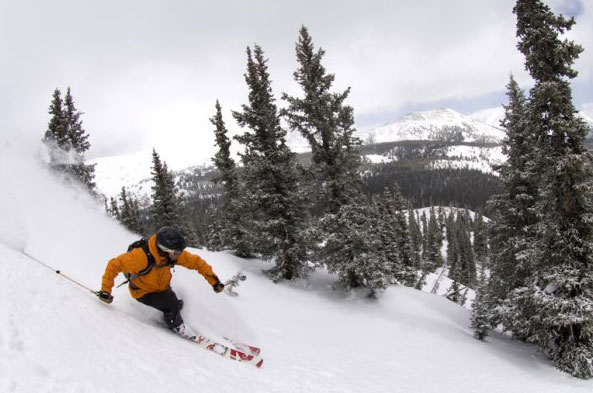 A skier wearing a yellow jacket skis down a snowy slope with a view of snow-covered mountain peaks in the distance. 
