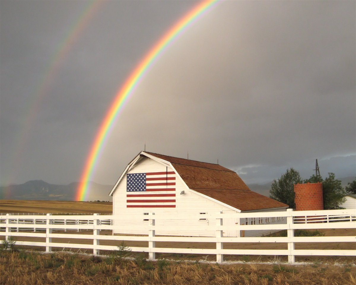 A rainbow protrudes from the grey, stormy sky above a white barn with an American flag painted on it. 