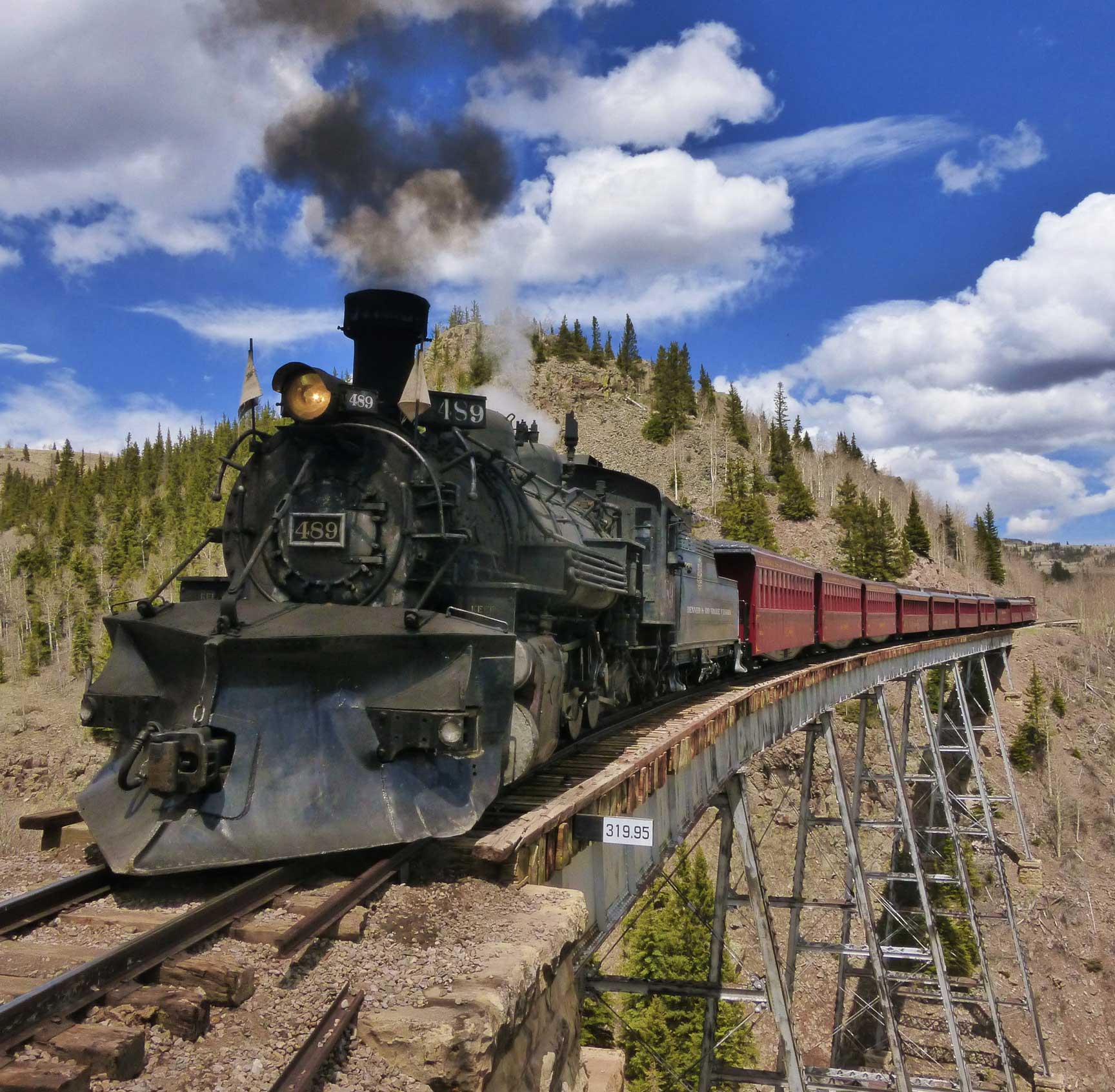 Crossing a trestle bridge on a blue-skied day with bright-white clouds, the Cumbres & Toltec Scenic Railroad train makes its way above a rocky valley.