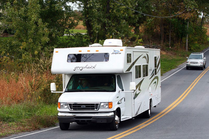A white RV drives down a leafy road