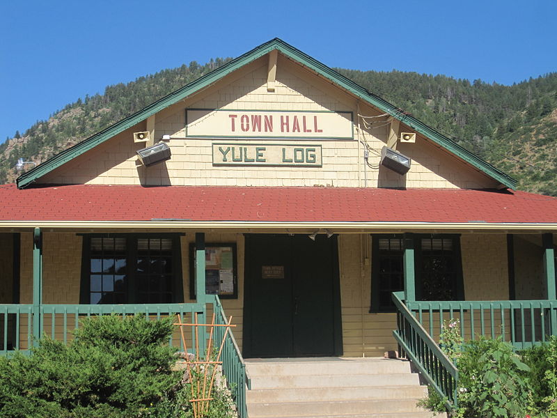 A yellow wood building with a red roof with green trim has a sign on it that says "Town Hall Yule Log." In the background evergreen trees atop a hill meet a blue sky.