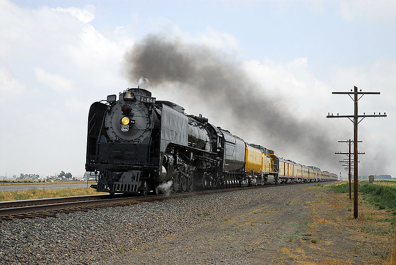 A train with black engine cars and yellow cars in the back steams across a train track with a gravel path to the left with telephone polls. The sky is light-blue with white clouds. 