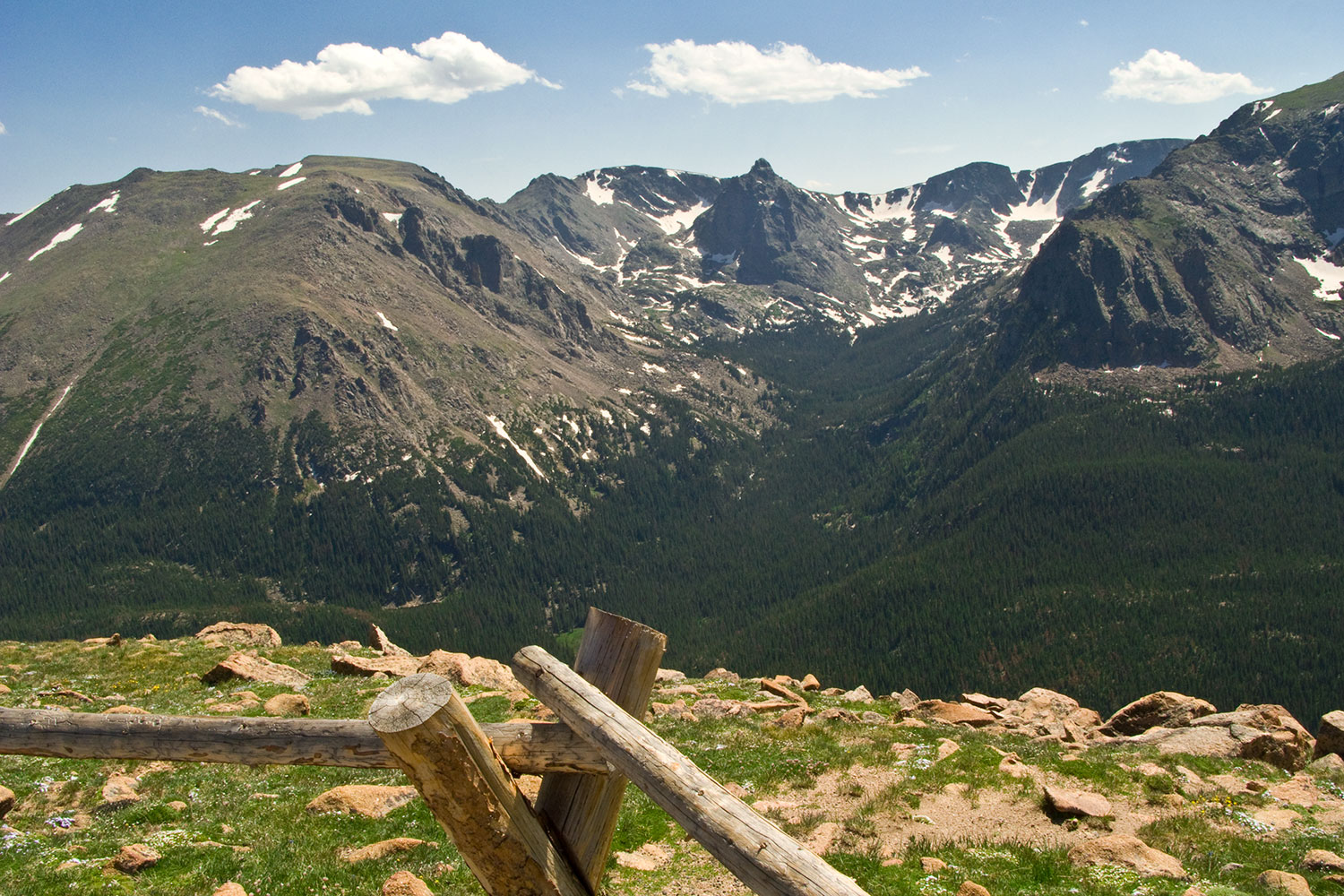 A view taken from an observation point of the Rocky Mountains with some of the stone peaks covered in snow. The sky is a hazy blue with three white clouds. There is green grass with rocks in front of the wooden fence at the observation point.