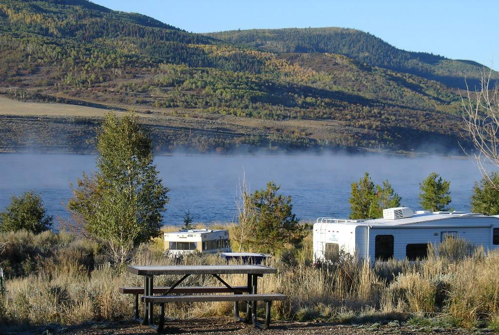 Two white RVs camp lakeside at Stagecoach State Park . There's a picnic bench in the foreground and a lake in the background with rolling, green-covered hills.