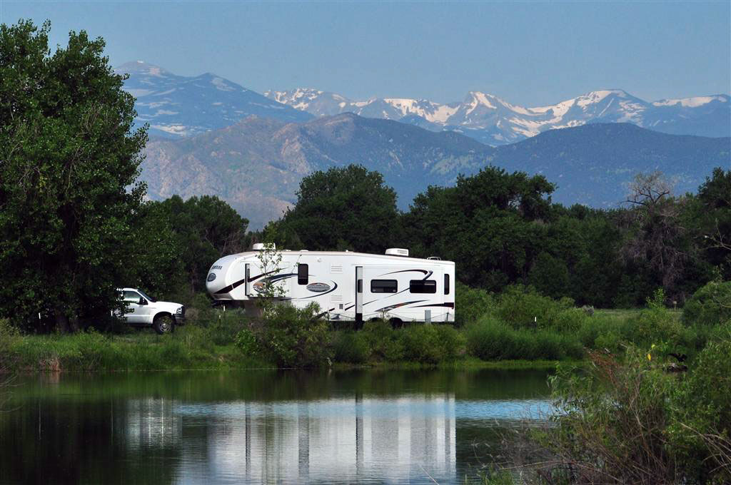 A white RV is parked next to a lake. Behind it is a lush grove of trees and snowtopped mountan peaks