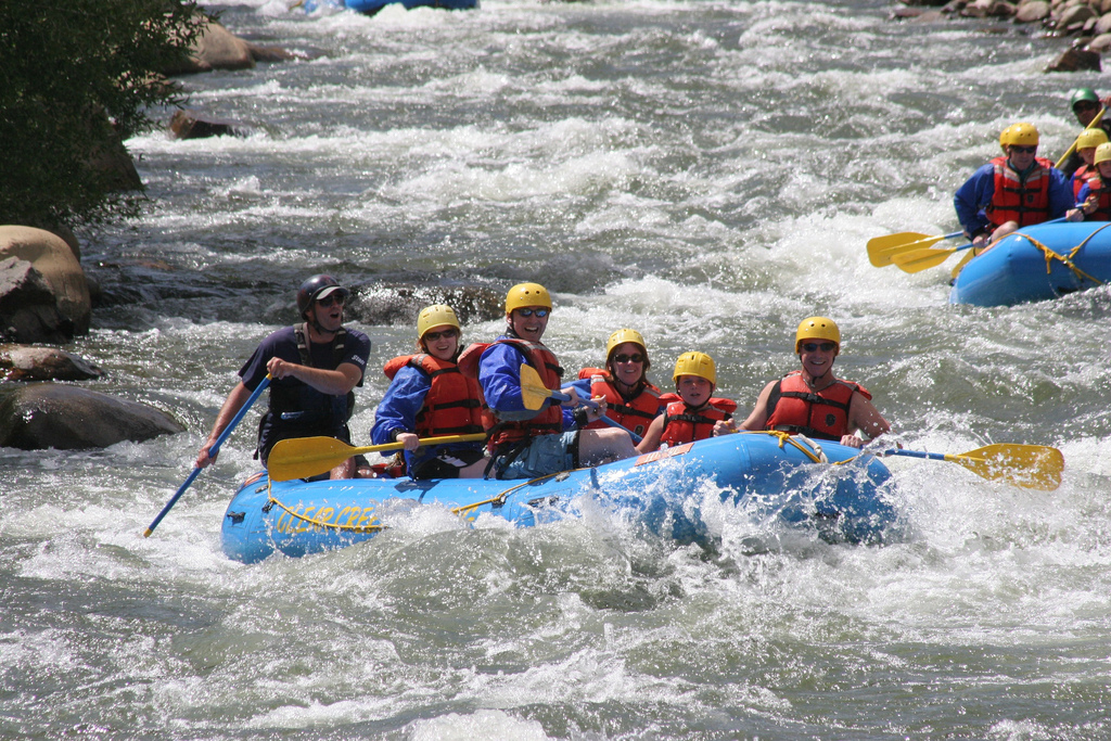 Eight people in red lifejackets inside a blue raft paddle through white rapids; a guide at the back tries to steer the raft through the water