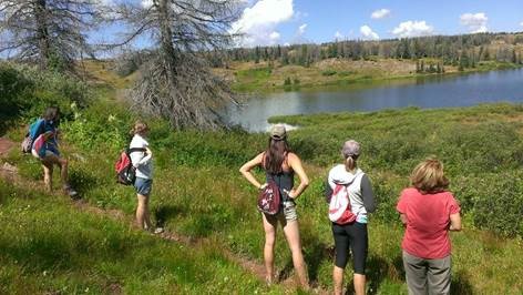 A group of hikers pause on a narrow dirt track that cuts through grassy foothills near Alamosa, Colorado. They admire the tranquil, sky-reflectinglake nearby.