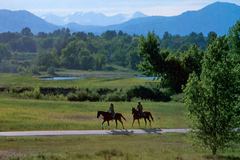 Two people riding horses along the South Platte River in South Platte Park. The grass is green and the trees in the distance have green leafs. Hazy mountains rise up to meet a white sky.