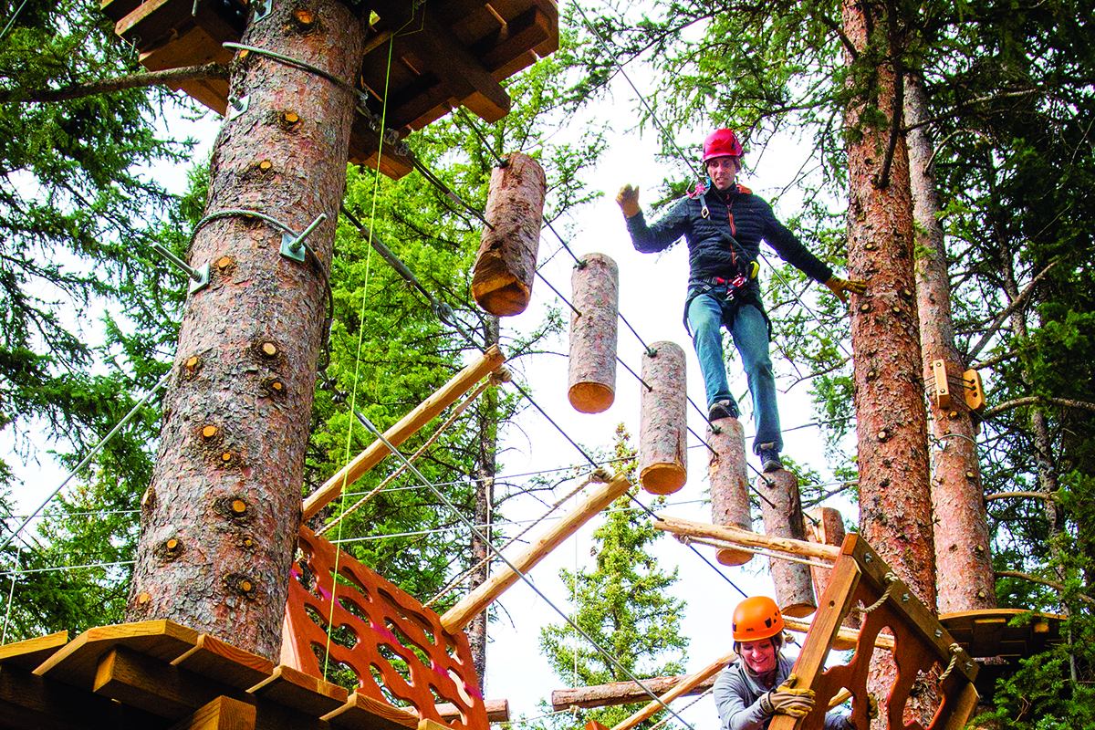 An adult wears a red helmet and is strapped into a safety harness as they explore a treetop playground and obstacle course among the pines at Lost Forest in Aspen, Colorado.