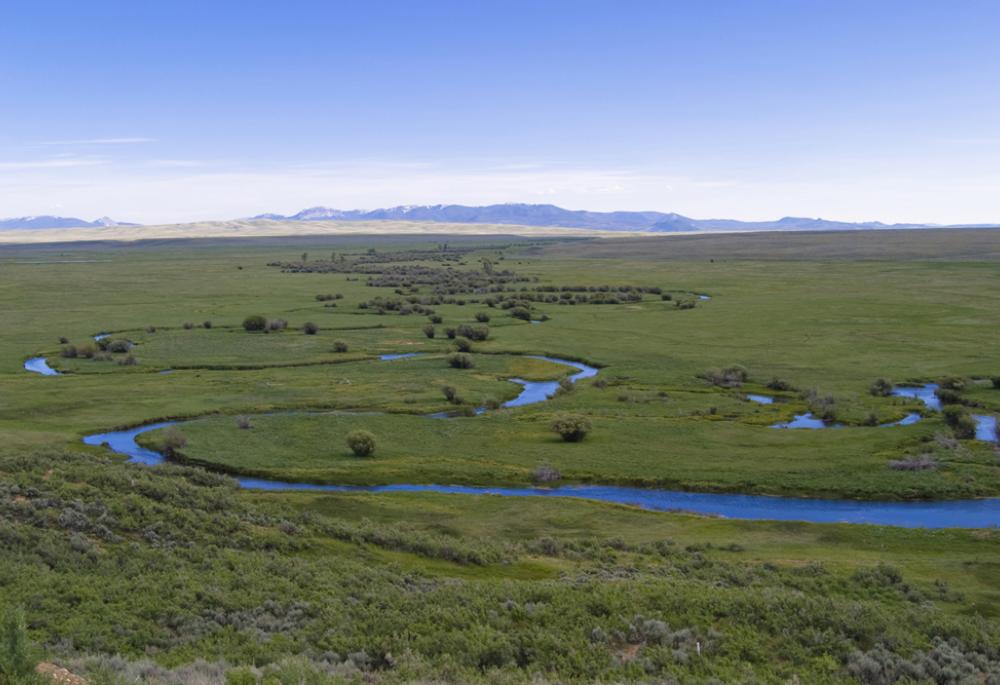 An aerial view of the snaking, blue Illinois River amongst green grasses. In the distance mountains rise up to meet a blue, hazy sky.