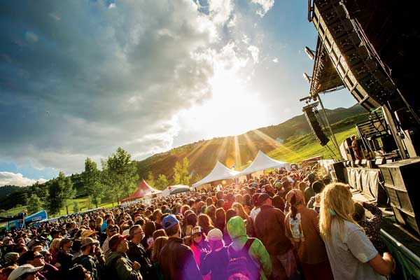 Bright rays of sunlight shine down through a cloudy sky onto the crowds at Snowmass Mammoth Fest. Around the large crowd a several tented booths and a speaker-loaded stage.