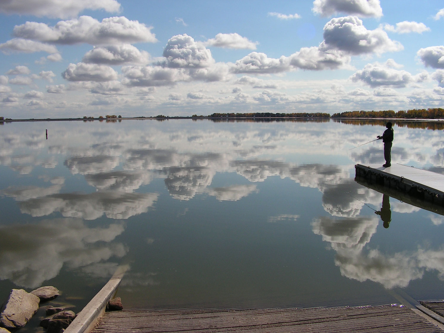 A solo fisher stands on a dock on the right of a glassy lake that is reflecting the image of a light-blue sky with many clouds. 