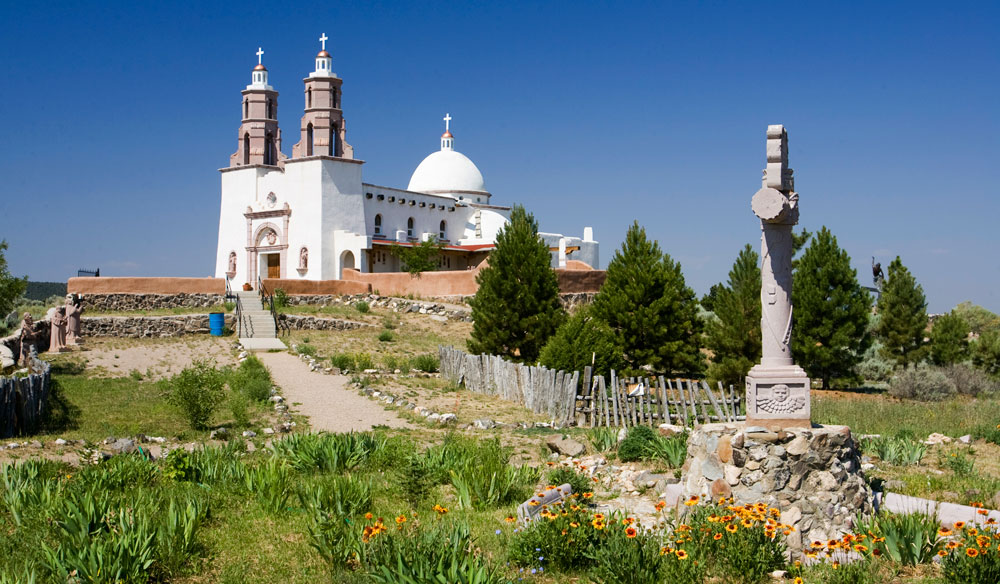 On a hill above a green-grass field with colorful wildflowers sits a bright-white church with two towers and a dome. The sky is blue, there are evergreen trees in the middle of the photo and a path leads through the grass to the Shrine of the Stations of the Cross building.