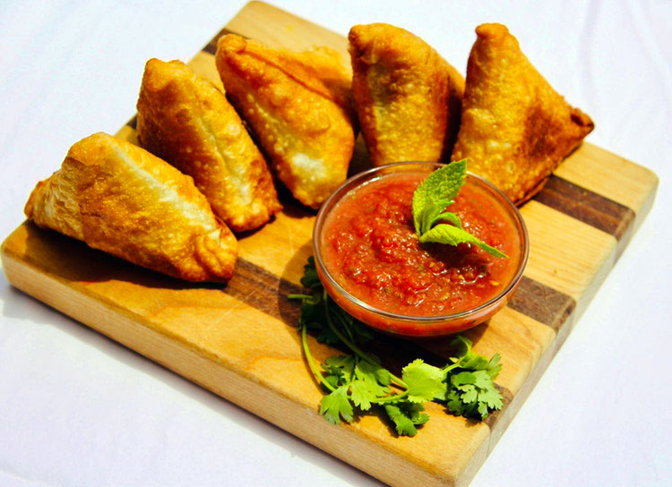 Five crispy samosas rest on a wooden serving board at Susan's Samosas in Colorado. They are served with a small glass dish of a tomato-based sauce.
