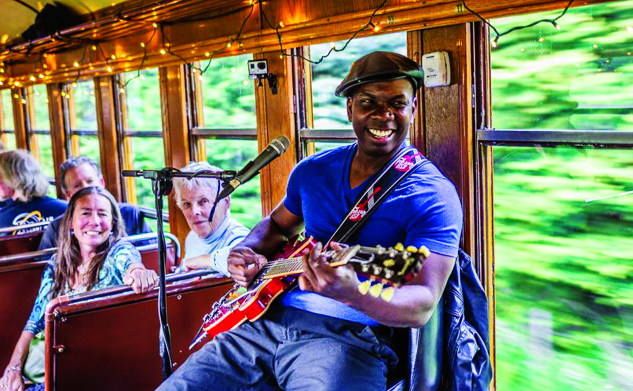 A musician smiles as they play a stop sign-red, electric guitar and sing to smiling passengers in a car of the Durango & Silverton Narrow Gauge Railroad’s Blues Train.