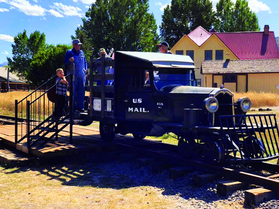 A child and several adults inspect an antique vehicle that appears to be equal parts train and automobile on a set of tracks at the Ridgway Railroad Museum in Colorado.