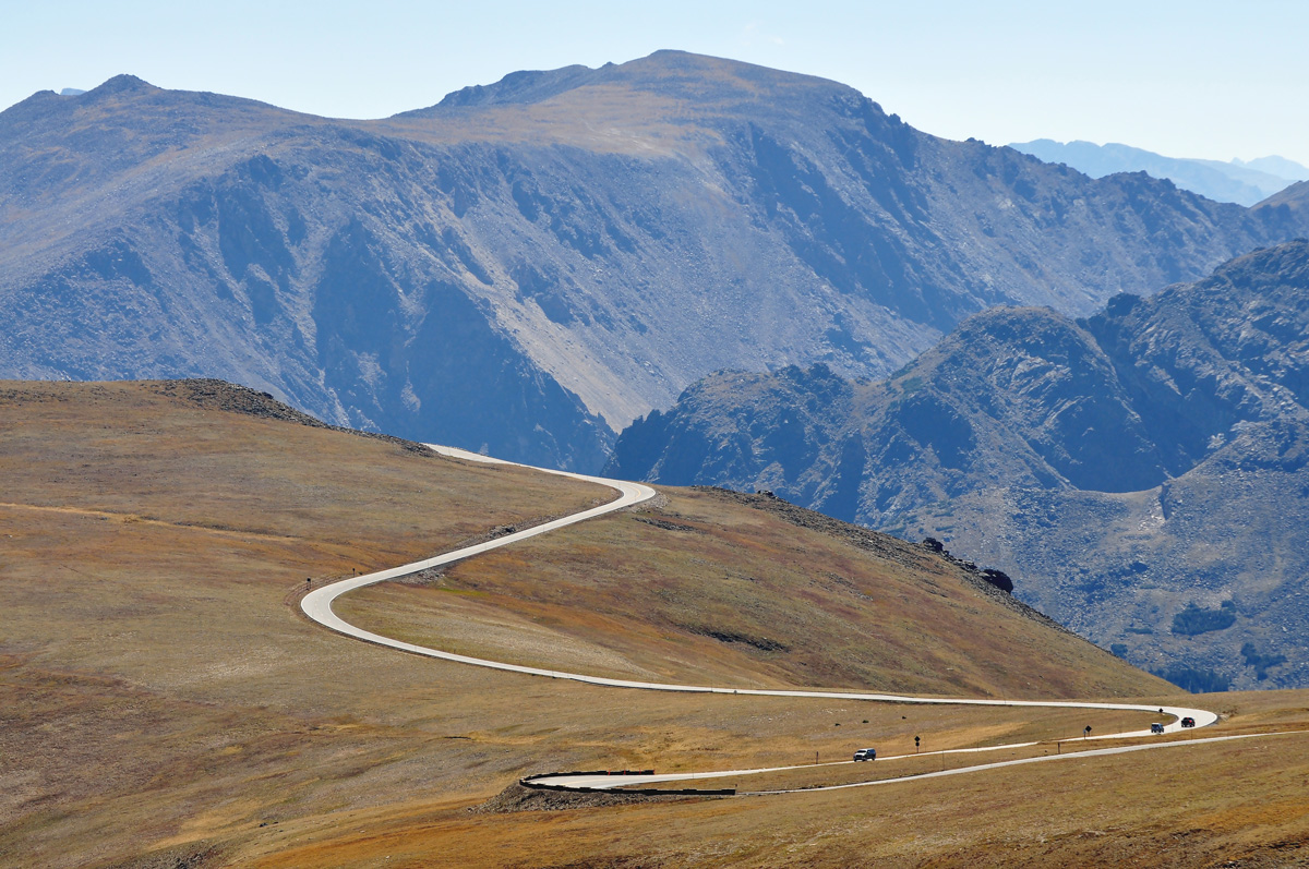 A handful of cars drive along the serpentine Trail Ridge Road in Rocky Mountain National Park in Colorado. In the background rugged mountain ridges seem to touch the sky.
