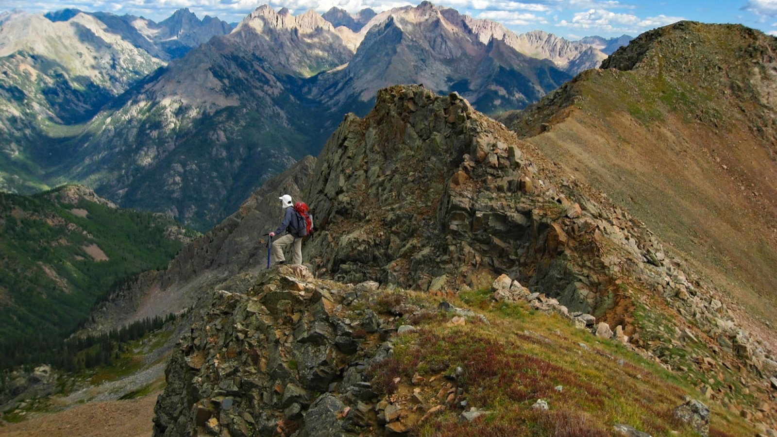 Someone in climbing gear and a helmet stands perched on top of the summit of Twilight Peaks in Weminuche Wilderness near Durango, Colorado. All around them are stunning, jagged peaks stretching as far as the eye can see under a mostly sunny sky.