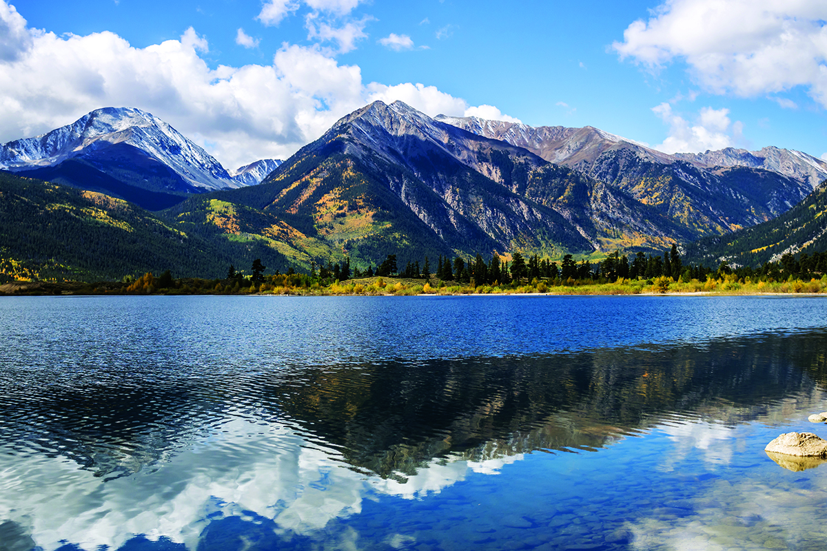 A lake reflects the image of a mountain range dusted with snow at the peaks and bursts of gold and orange fall foliage on an otherwise evergreen mountainside in Colorado.
