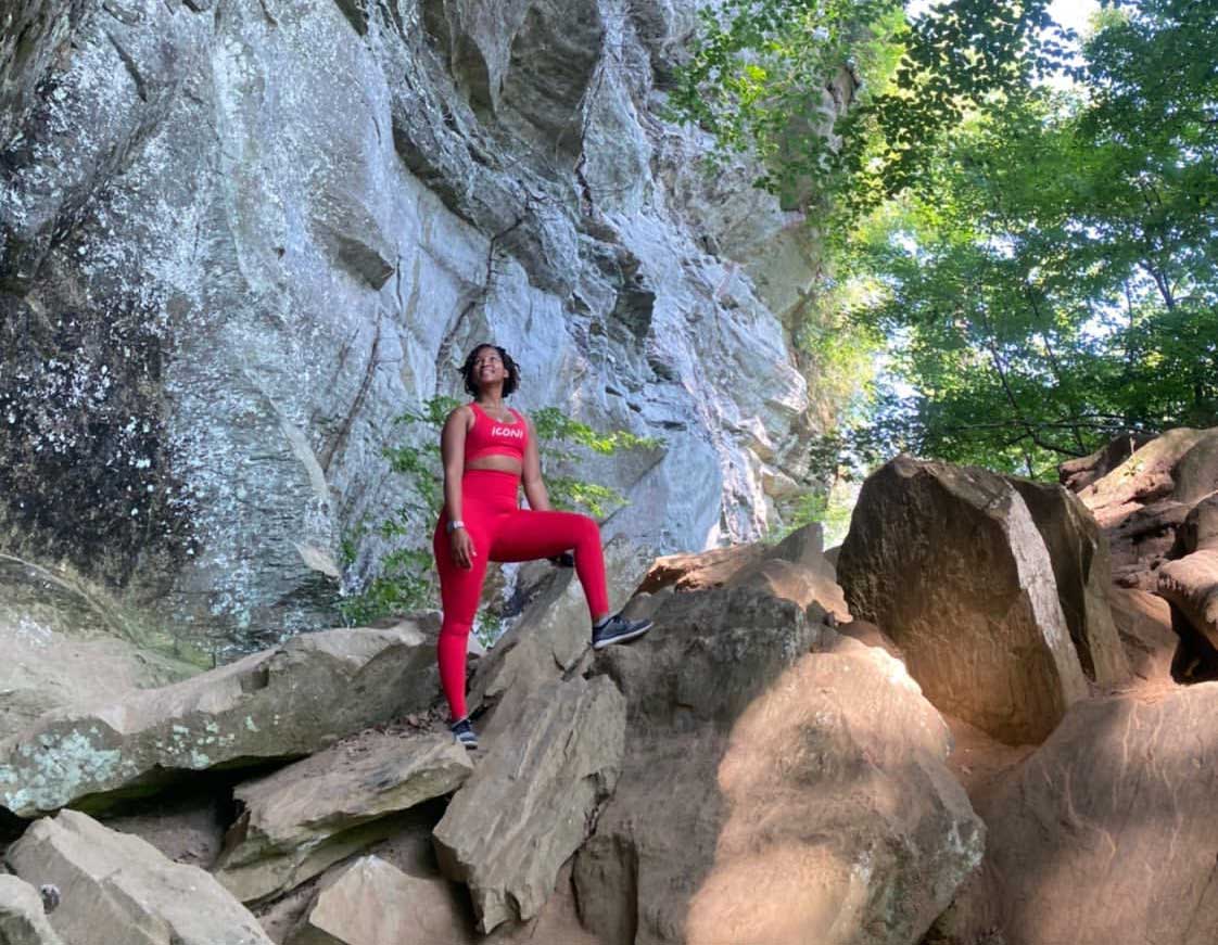 In Coloradoro, a model poses in a small rock field with a rock wall and trees around them. They wear a bright-pink activewear outfit.