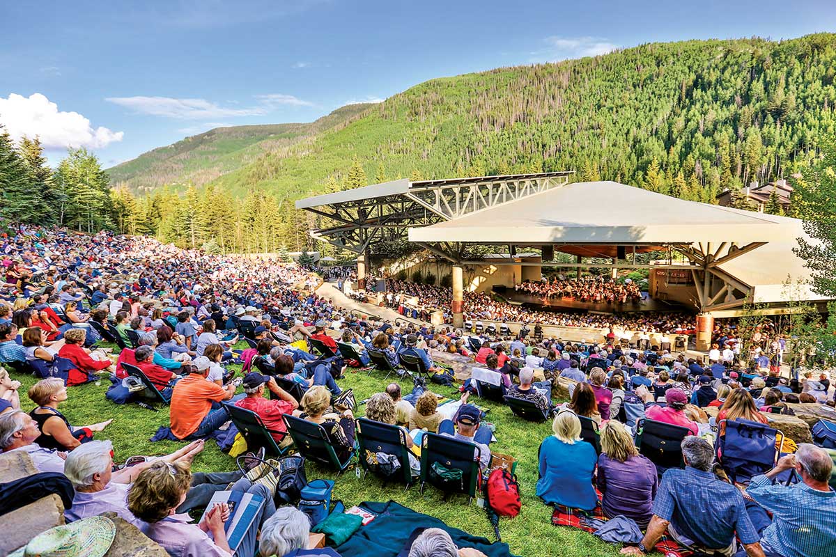 Groups of music fans sit on lawn chairs and blankets facing an amphitheater in Vail, Colorado, and listening to a performance as part of the Bravo! Vail Valley Music Festival.