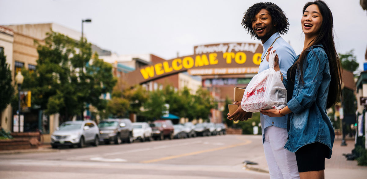 Two people, one holding a takeout container, both wearing blue stand on the sidewalk looking up the street. In the background the "Welcome to Golden" arch sits with cars parked parallel.