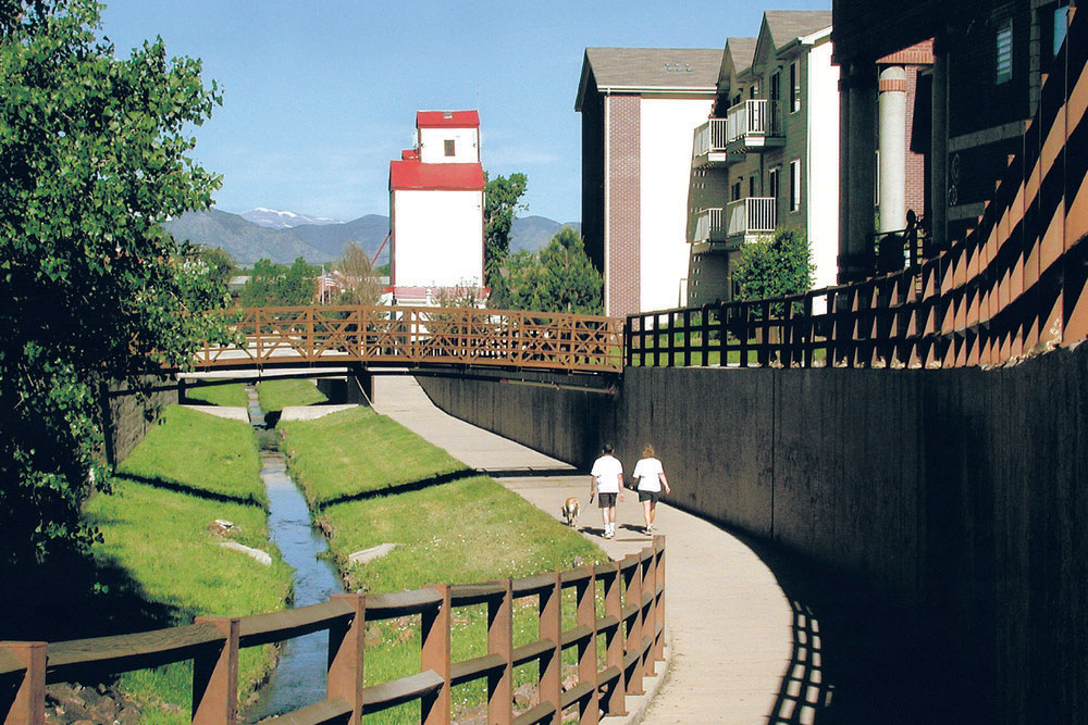 A couple walks a dog along the Littles Creek path with green banks of a creek to the left and a cement wall topped with a brown fence to the right. In the distance a pedestrian bridge and tall white building with a red roof meet the blue sky. 