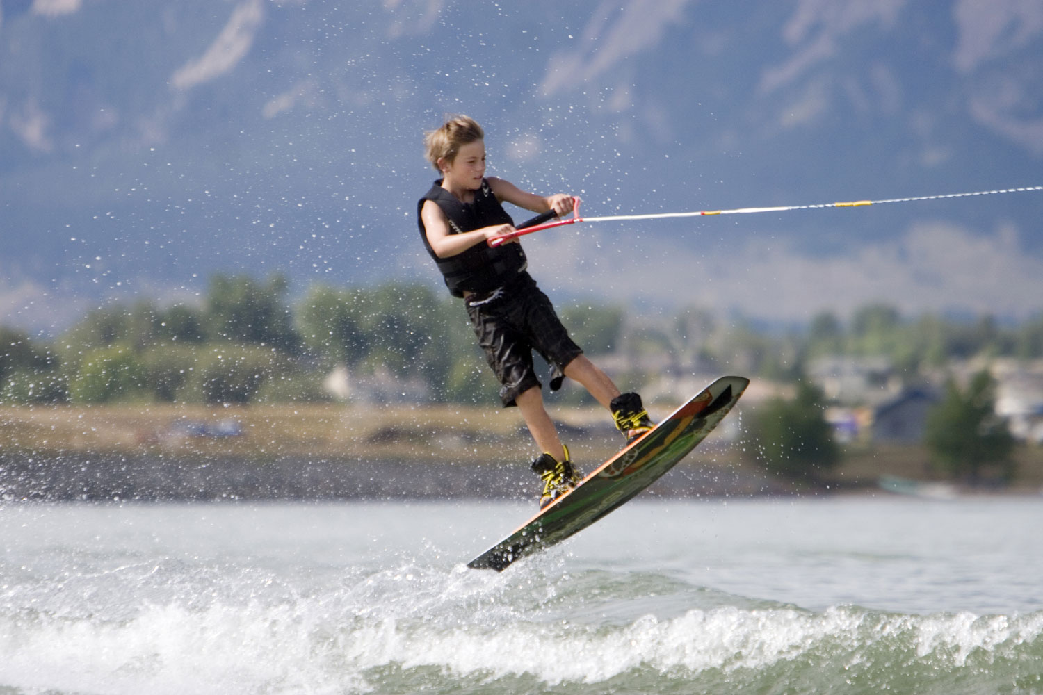 A boy on a wake board being towed by a boat launches into the air over some rapids; he wears a life vest