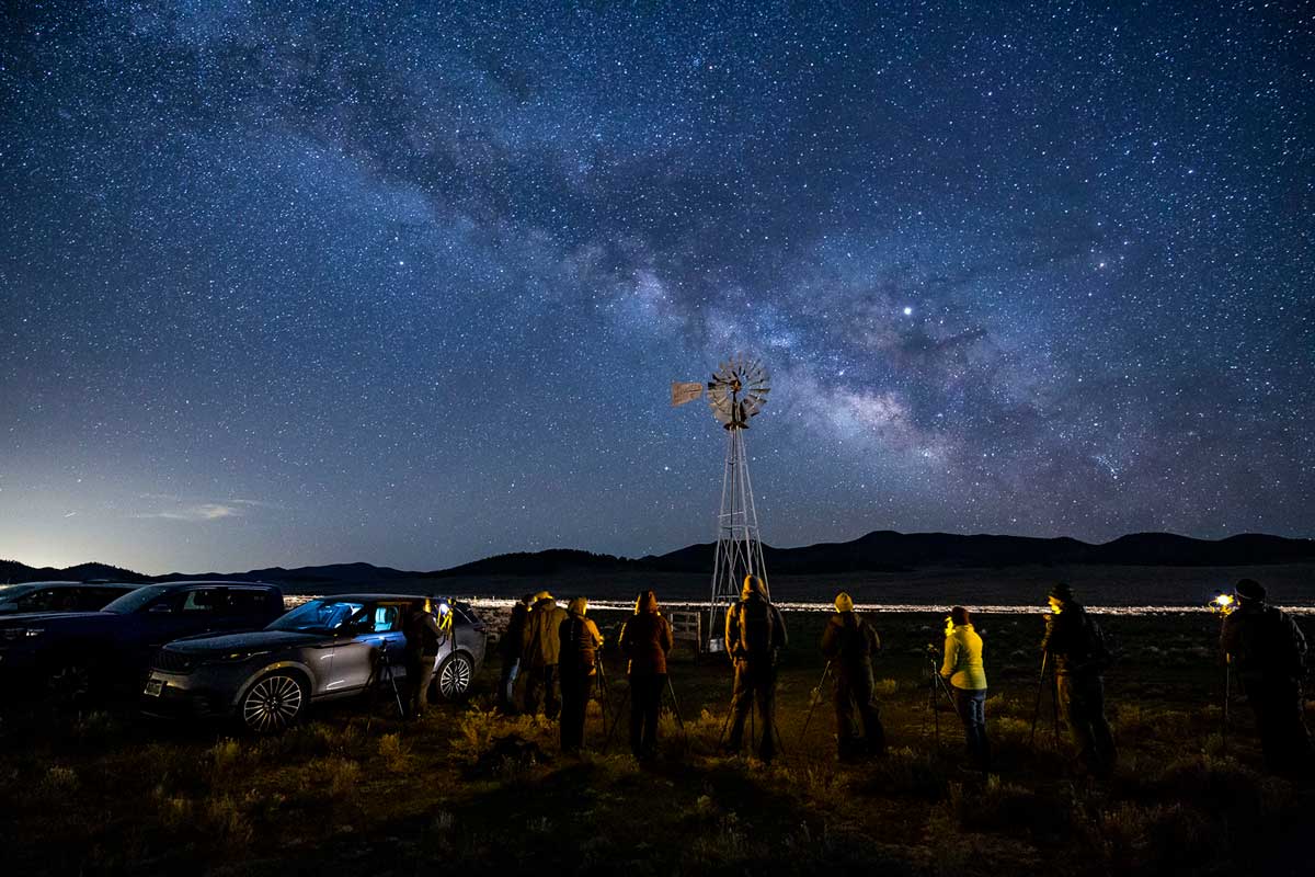 Strargazers stand looking up at the night sky with the Milky Way and tons of stars. There's an old-school windmill and two parked cars as well.