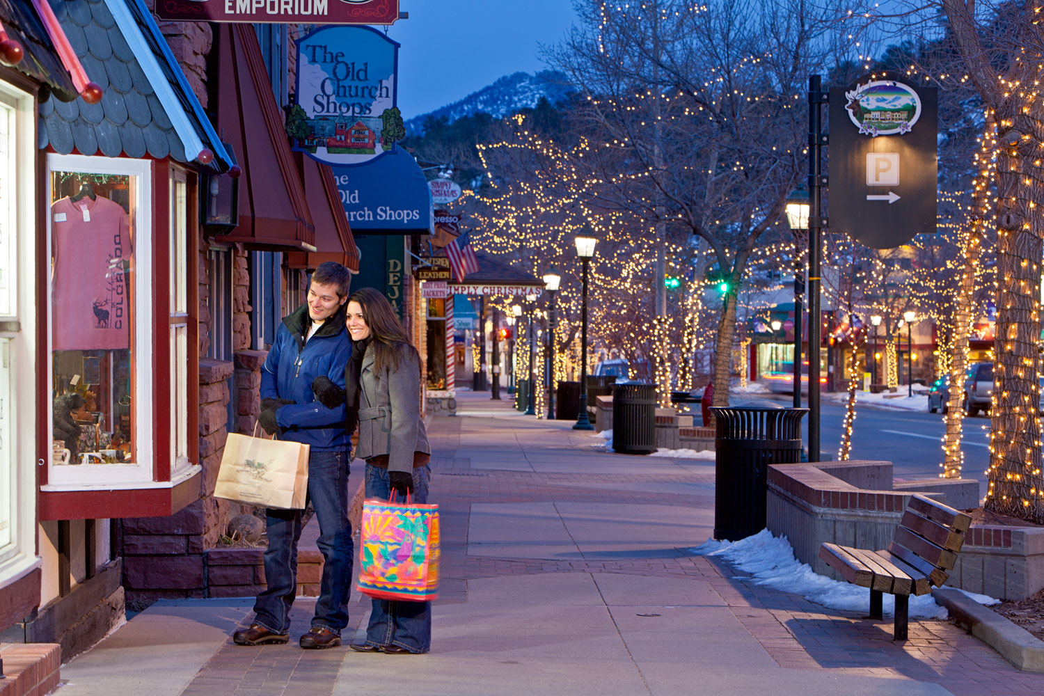 String light-lit trees line the wide sidewalks of Estes Park, Colorado. A couple pauses with their shopping bags and looks in a boutique.