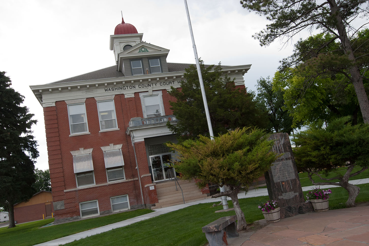 Front of the red-brick, two story Washington County Courthouse in downtown Akron, Colorado. The trees and grass are green. The cupola at the top of the building is red. 