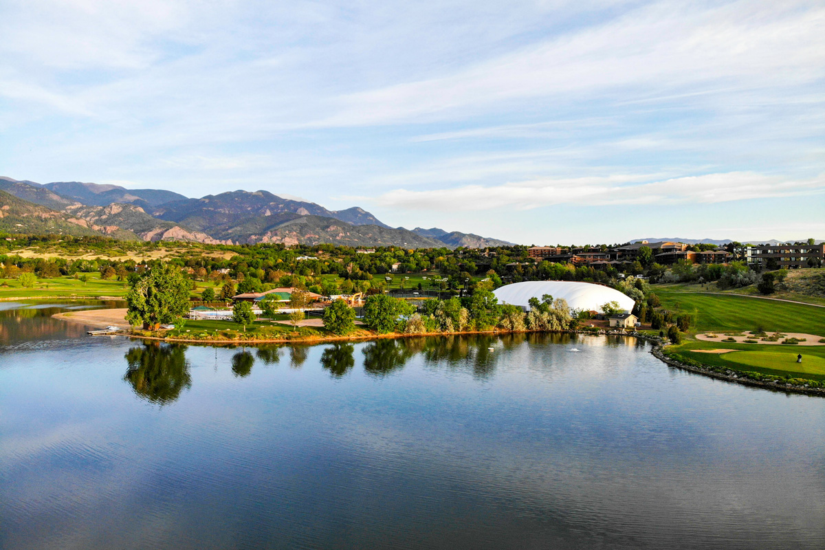 Bluebird skies with wisps of clouds are reflected in the calm waters near Cheyenne Mountain Resort. There is a golf course by the edge of the water and the resort area looks lush with many mature trees.